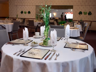 Round dining tables in a decorated dining room set with white tablecloths, place settings, folded napkins and green centerpieces.