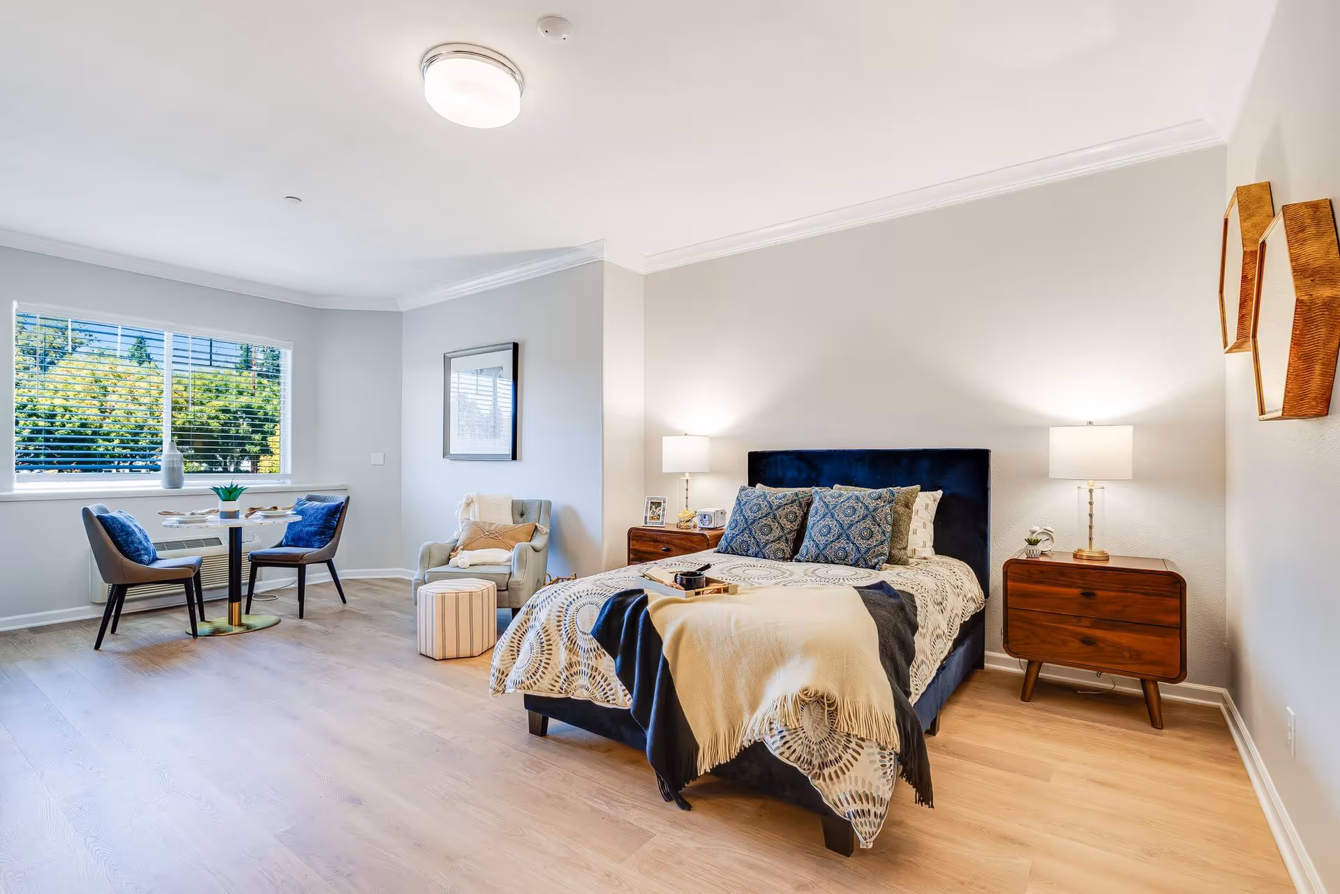 A bright and spacious bedroom with a large bed featuring patterned bedding and multiple pillows. There are two wooden nightstands with lamps on either side of the bed. To the left, there is a cozy sitting area with a cushioned armchair, a small ottoman, and a round table with two chairs near a window with blinds. The room has light-colored walls and wood flooring, with decorative wall art on the right side.