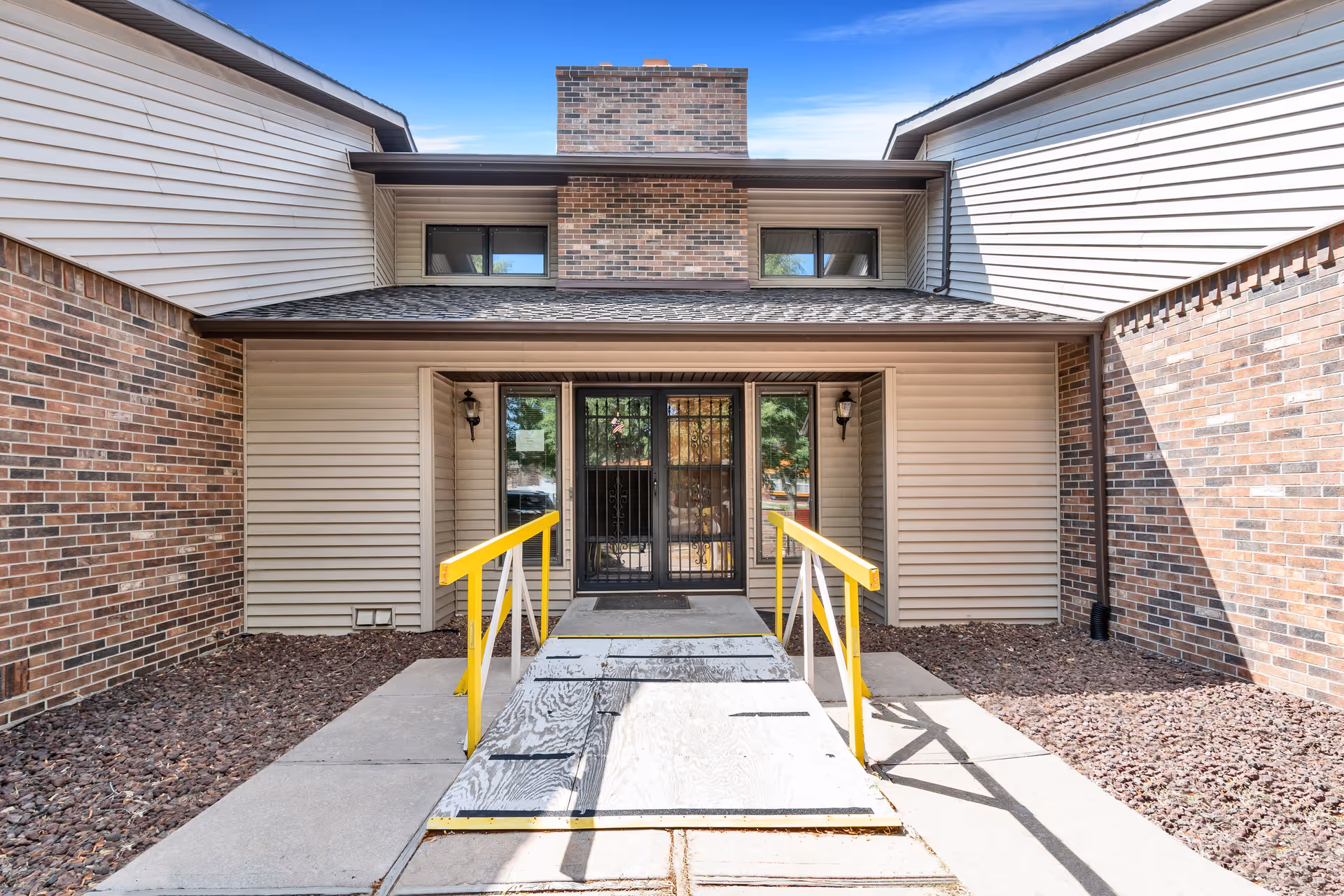 Entrance to a building with a wooden ramp featuring yellow handrails leading up to double glass doors with metal security bars. The building exterior has beige siding and brick walls, with a chimney above the entrance and two small windows on either side of the chimney. The sky is clear and blue.