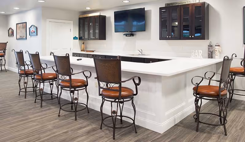 Bright communal kitchen/bar area with a white island countertop, mounted TV, cabinets, and several high-backed bar stools.
