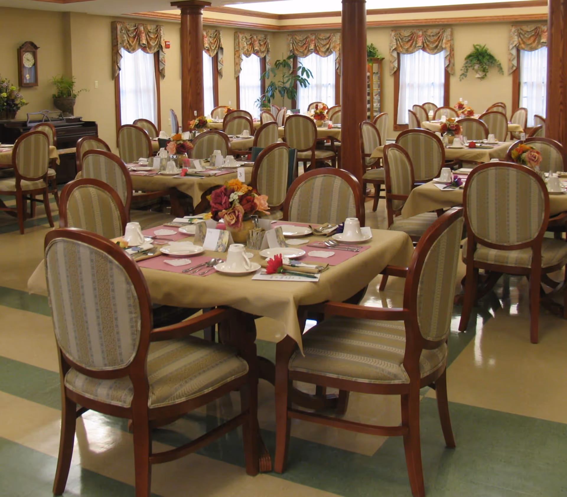 Dining room with multiple tables set with place settings and upholstered chairs in a senior living facility.