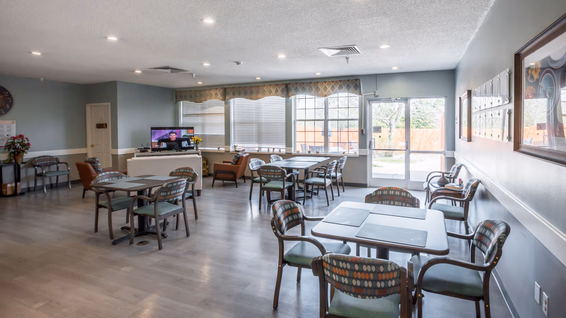 A spacious common area in a senior living facility with several tables and chairs arranged for dining or socializing. The room has large windows with blinds, a TV on a stand, and a door leading outside. The walls are painted light blue with framed artwork and a clock. The floor is light wood, and the room is well-lit with recessed ceiling lights.