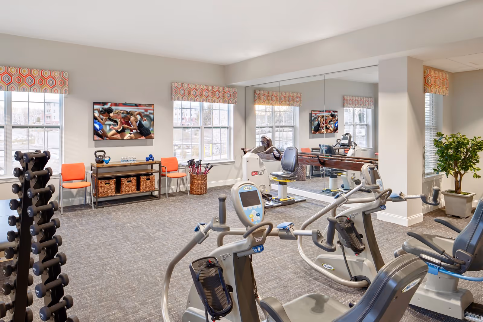 A bright fitness room with exercise equipment including stationary bikes and a weight rack. The room has large windows with patterned valances, a wall-mounted TV showing a running race, a console table with baskets and water bottles, and a large mirror covering one wall. There are two orange chairs and a basket with walking sticks near the windows, and a potted plant in the corner.