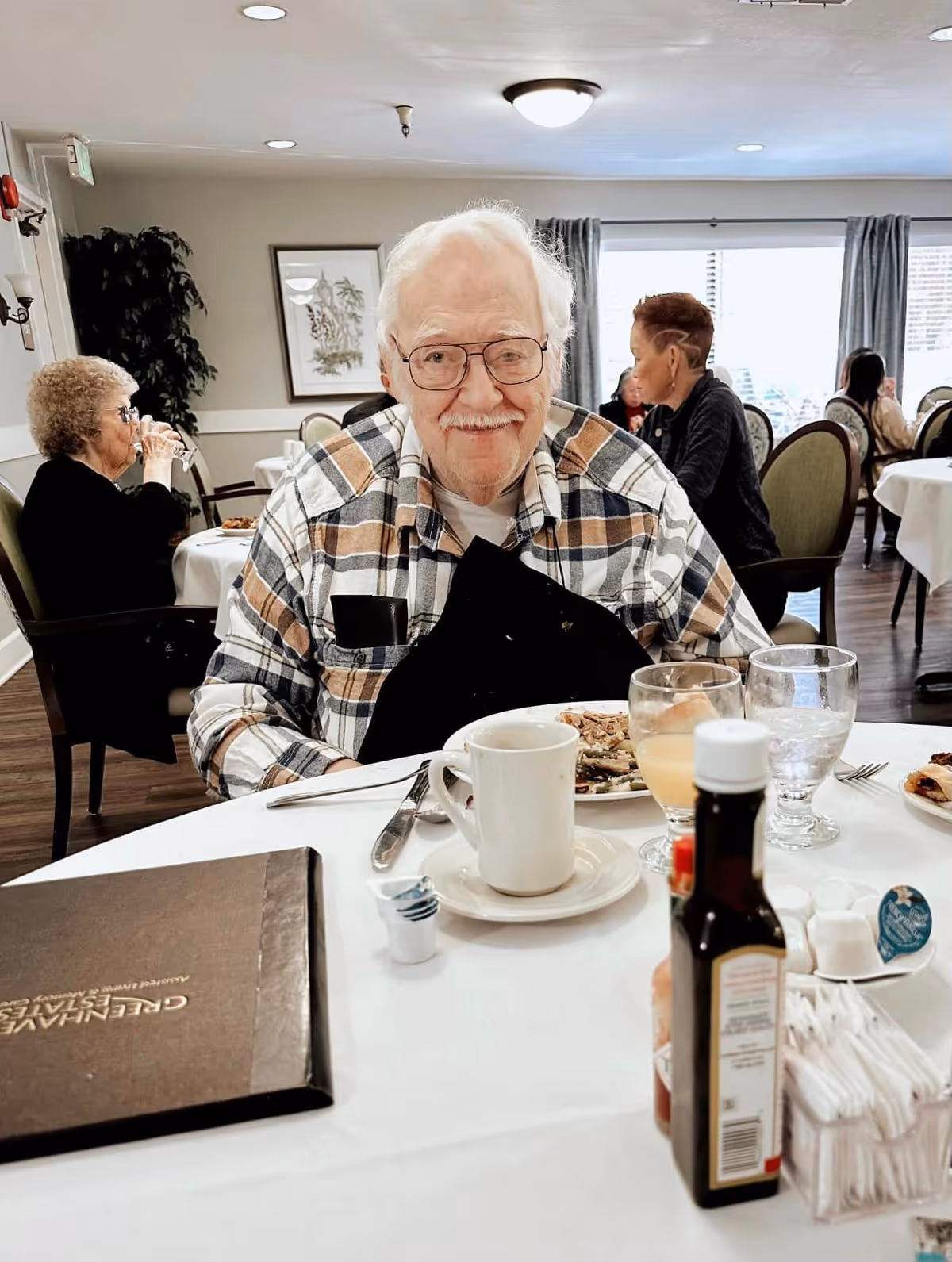 An elderly man in glasses and a plaid shirt smiles while seated at a dining table in a communal dining room.