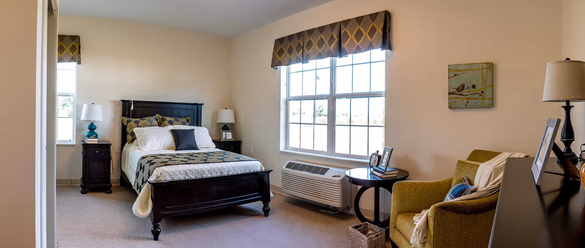 A well-lit bedroom with a large window covered by a valance, a dark wooden bed with patterned bedding, two matching nightstands with lamps, a green armchair with a throw blanket, a round side table with picture frames, and a wall-mounted air conditioning unit beneath the window.