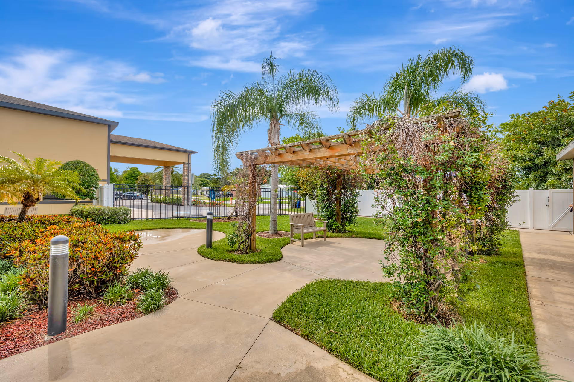 Courtyard with a vine-covered wooden pergola, a bench, palm trees, walkways, and landscaped greenery outside the building.