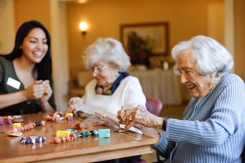 Two elderly women and a younger woman sitting around a wooden table engaged in a craft activity with colorful clay pieces in a warmly lit room.