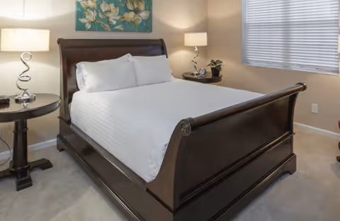 A neatly made bed with white linens and pillows in a bedroom. The bed has a dark wooden frame with a sleigh design. There are two matching dark wooden nightstands on either side of the bed, each with a modern lamp and decorative items. A window with closed blinds is on the right wall, and a floral painting hangs above the bed.