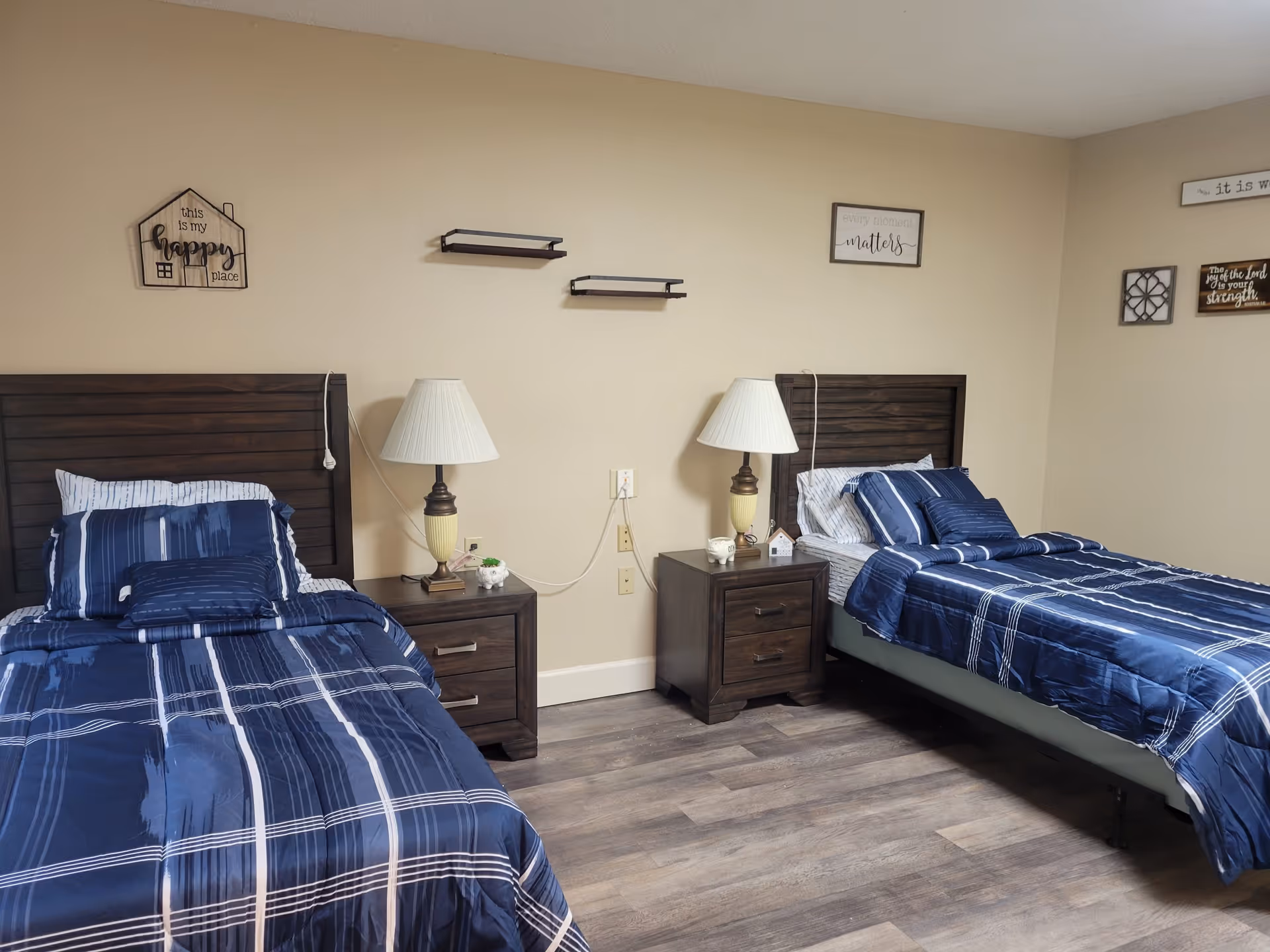 A bedroom with two twin beds dressed in blue plaid bedding, a shared nightstand with lamps, and wall decor.