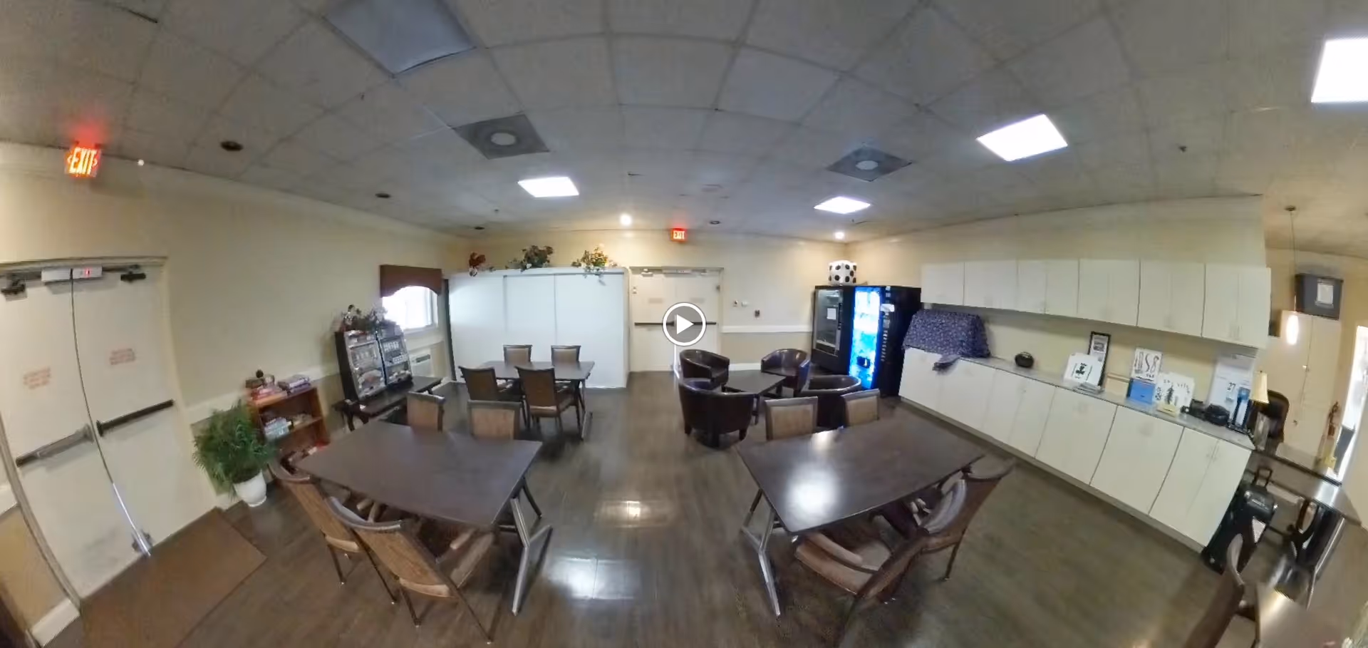 Interior view of a common area in The Colony Club Sunrise featuring several tables with chairs, a vending machine, a small seating area with armchairs, cabinets along one wall, and double exit doors. The room has a tiled ceiling with fluorescent lighting and wood flooring.