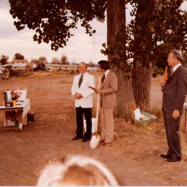 Three men in suits stand outdoors by a large tree during a ceremonial groundbreaking, one holding a shovel.