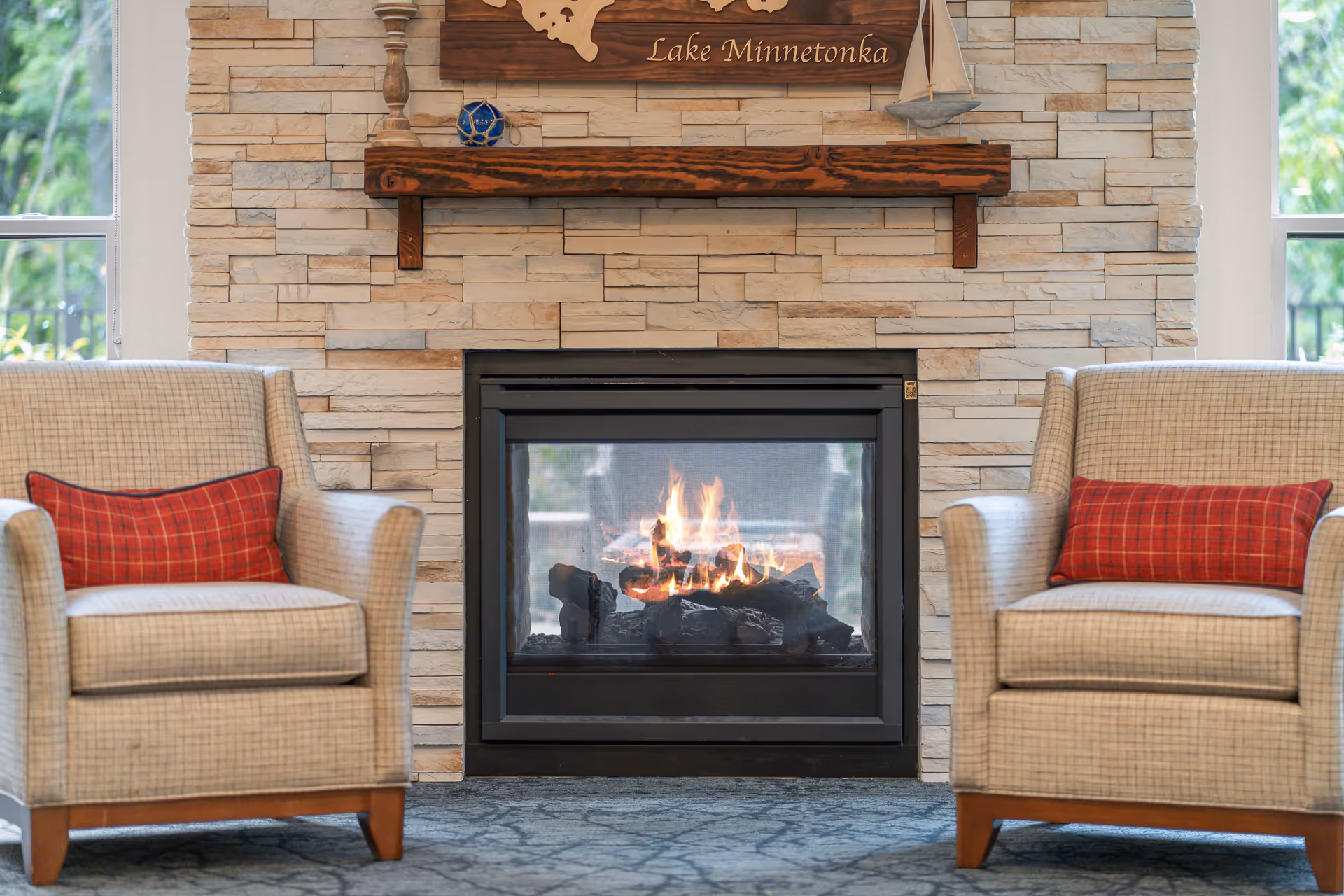 Two beige armchairs with red plaid cushions facing a lit fireplace set in a stone wall with a wooden mantel. Above the mantel is a wooden sign that reads 'Lake Minnetonka'.