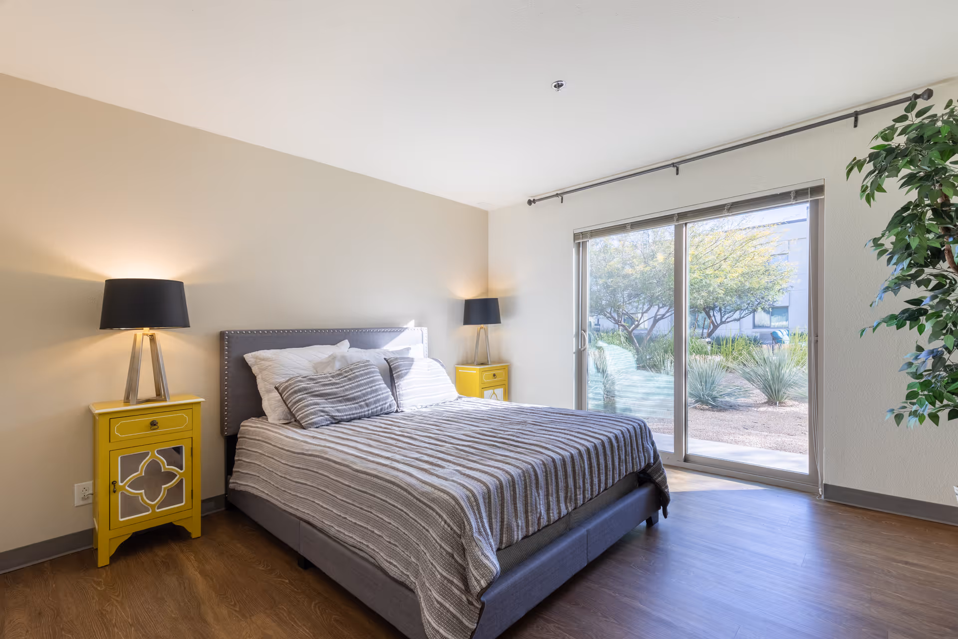 A bright bedroom with a large bed featuring a gray upholstered headboard and striped bedding. There are two yellow nightstands on either side of the bed, each with a modern black lamp. A large sliding glass door lets in natural light and offers a view of trees and plants outside. A green potted plant is visible on the right side of the room.