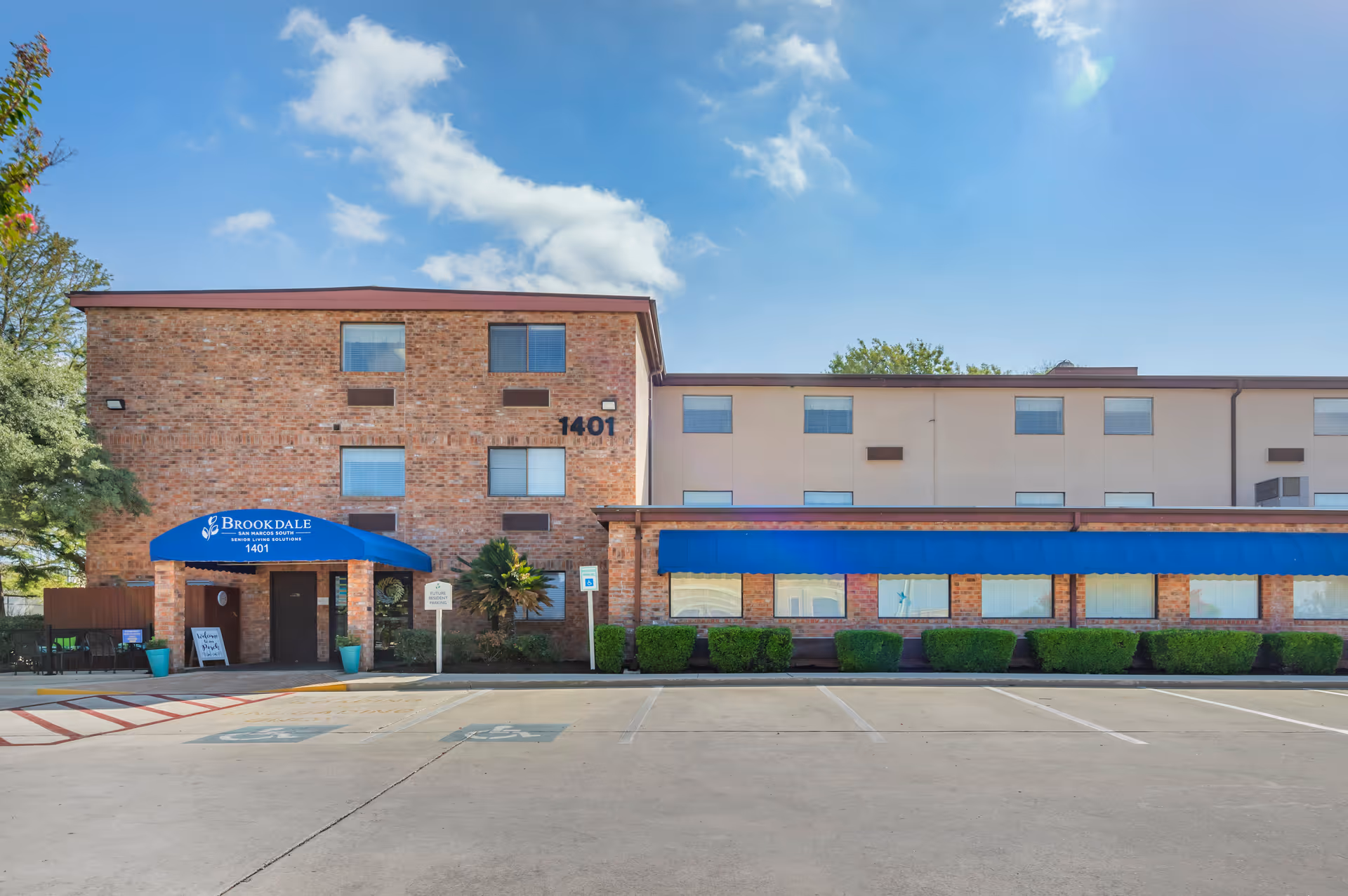 Exterior view of Brookdale San Marcos South senior living facility showing a brick and beige building with multiple windows, a blue awning over the entrance, and a parking lot with handicap spaces in front.