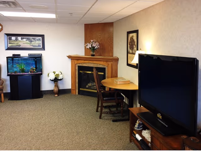 A cozy living room area featuring a large flat-screen TV on a wooden stand, a round wooden table with a chair, a fireplace with a wooden mantle decorated with a vase of flowers, an aquarium on a stand, a framed picture on the wall, and a vase with white flowers on the floor.