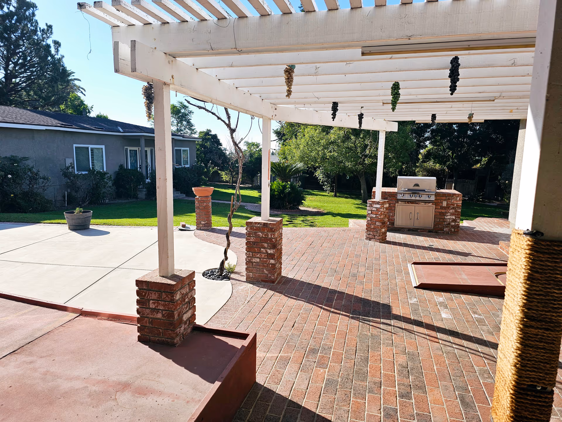 Outdoor patio area with a white pergola supported by brick pillars. There are bunches of artificial grapes hanging from the pergola. The patio has a brick floor and includes a built-in stainless steel barbecue grill. Surrounding the patio is a green lawn with trees and shrubs, and a single-story building with windows and a door is visible in the background.