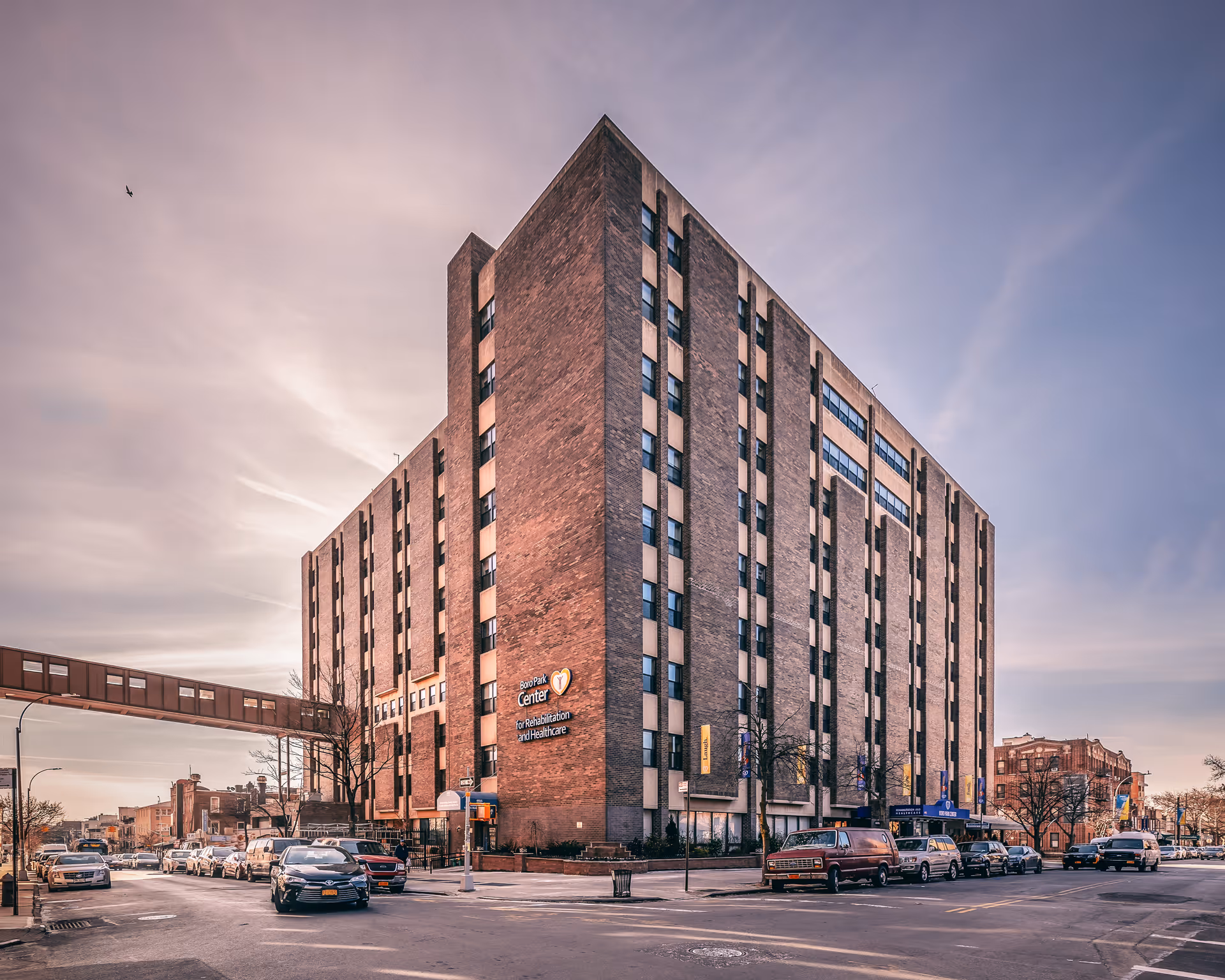Exterior view of a large multi-story brick building named Boro Park Center for Rehabilitation and Healthcare, situated at a street corner with several parked cars and a pedestrian bridge connecting to another building.