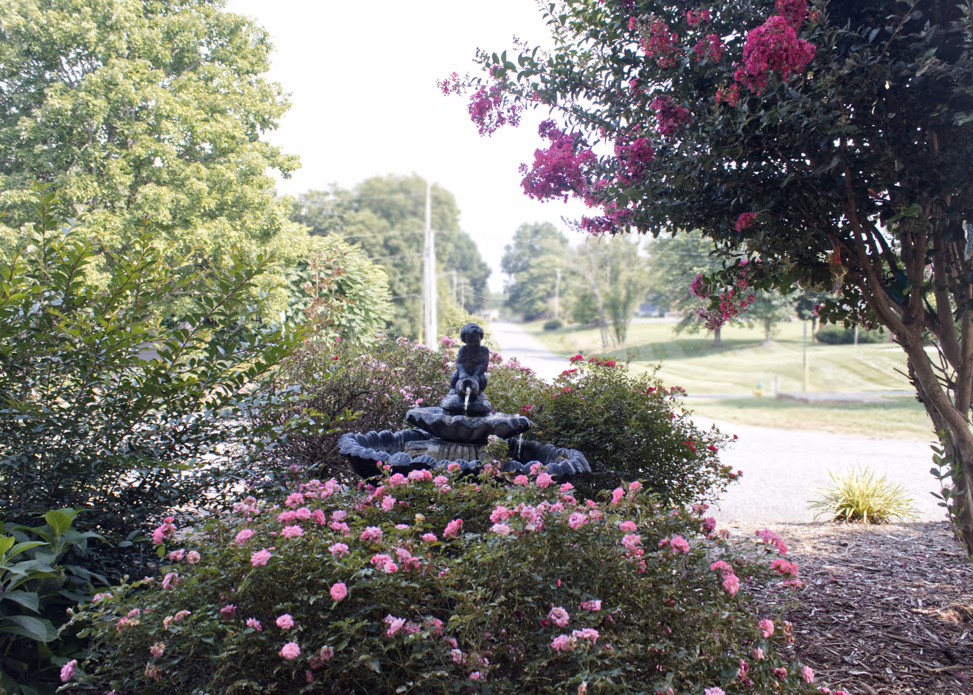 A garden area with a small decorative water fountain featuring a statue of a child holding a hose, surrounded by pink flowers and green bushes, with trees and a road in the background.