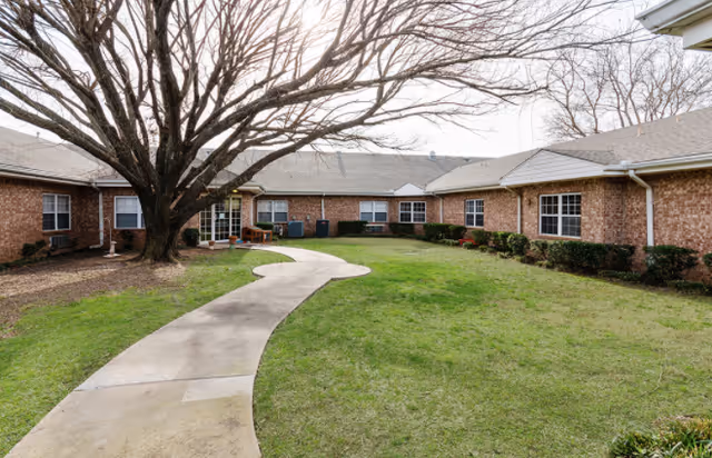Curved concrete path through a grassy courtyard with a large leafless tree and surrounding single-story brick building.