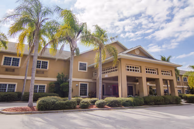 Exterior view of a two-story senior living facility building with beige walls and multiple windows. The entrance has a covered portico supported by columns. Several tall palm trees and well-maintained shrubs are in front of the building under a partly cloudy sky.