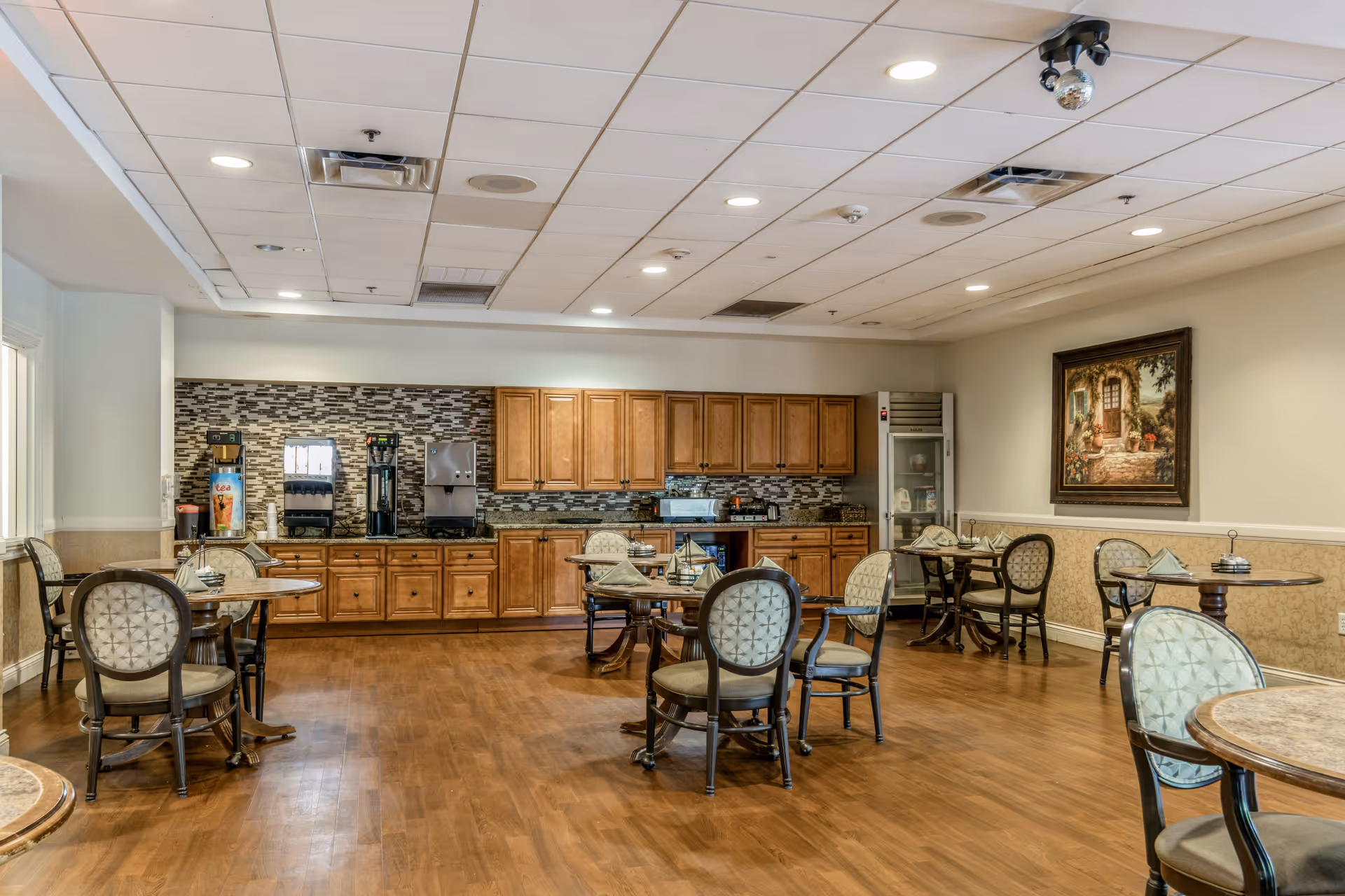 A dining area in a senior living facility with several round tables and cushioned chairs arranged around the room. The back wall features wooden cabinets, a beverage dispenser, and a small refrigerator. The floor is wooden, and the ceiling has recessed lighting. A framed painting hangs on the right wall.