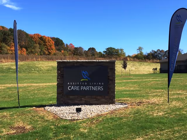 Stone entrance sign reading 'Assisted Living Care Partners' on a grassy lawn with tall flags and trees under a blue sky.