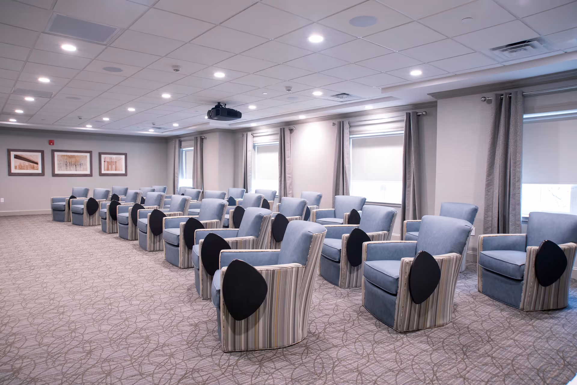 A spacious meeting or presentation room with multiple rows of blue upholstered chairs, each equipped with a black foldable writing pad attached to the armrest. The room has a patterned carpet, several windows with gray curtains, framed artwork on the wall, and a ceiling-mounted projector.