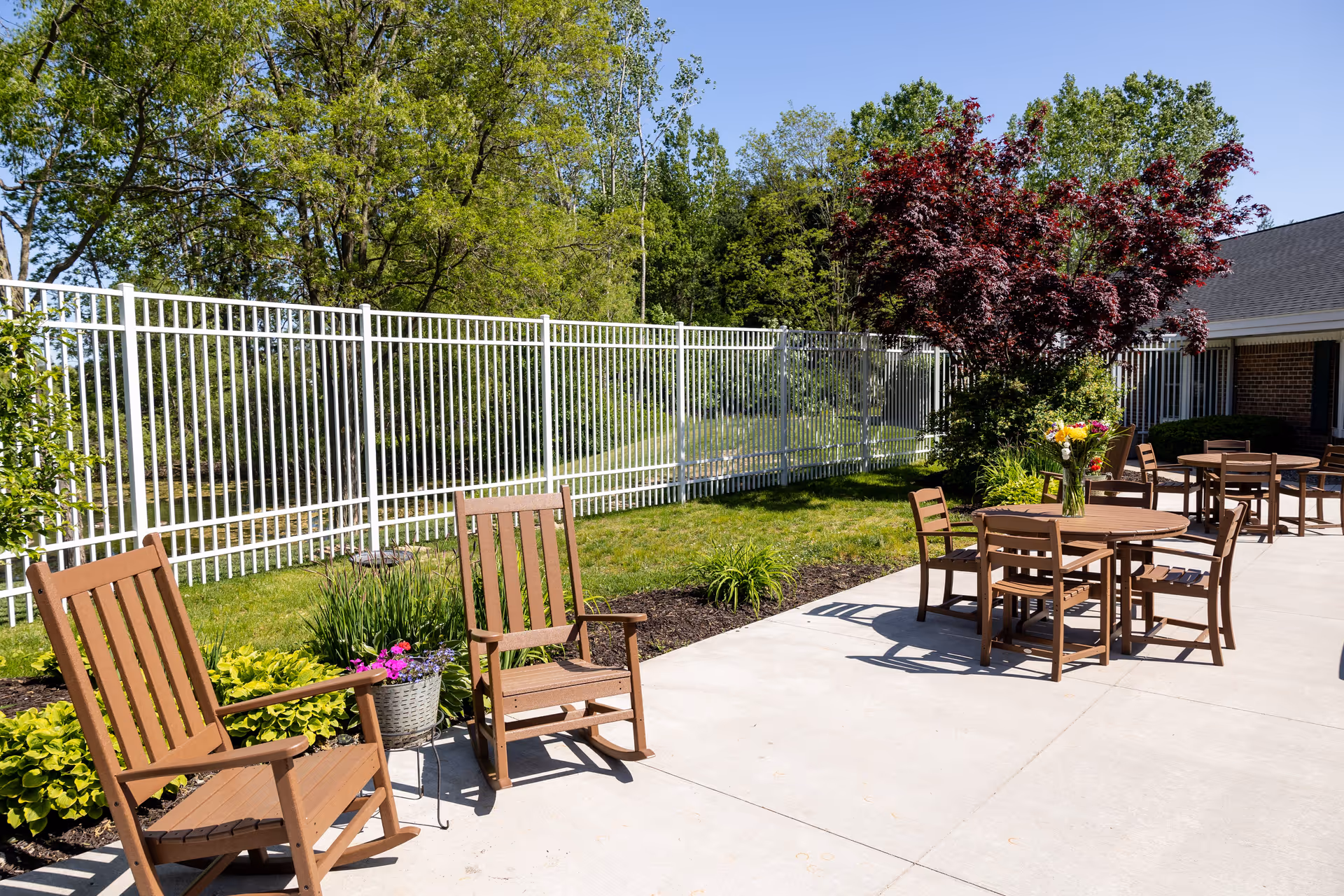 Sunny outdoor patio with wooden chairs and tables, a white metal fence, and landscaped plants.