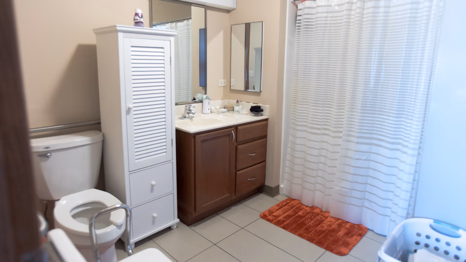 A clean bathroom with a white toilet, a white storage cabinet, a wooden vanity with a sink and mirror above it, a shower with a white and gray striped curtain, and a red bath mat on the tiled floor. A laundry basket is partially visible on the right side.