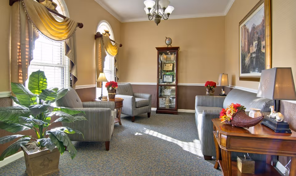 Sunny sitting room with upholstered chairs, side tables, potted plant and a glass display cabinet.