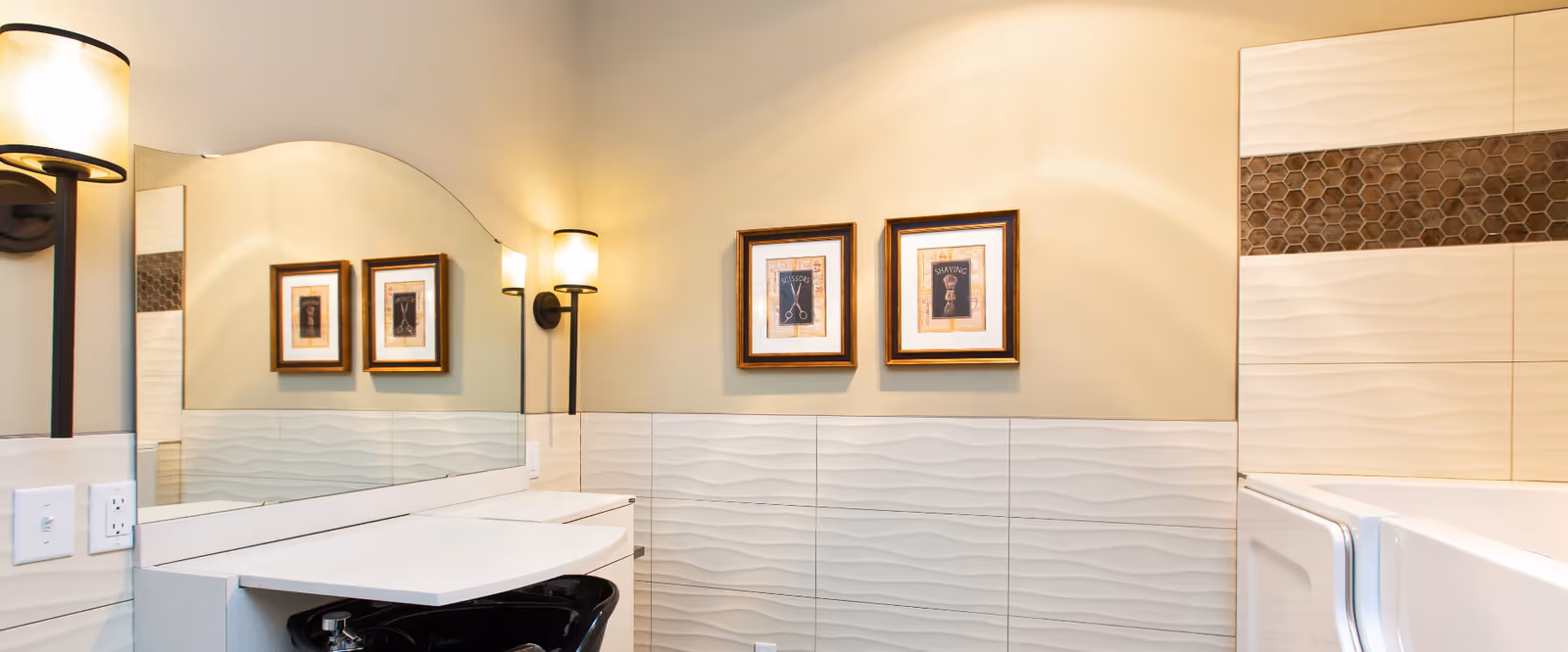 A modern bathroom interior featuring a large mirror with an arched top, two wall-mounted light fixtures with frosted glass shades, textured white wall tiles with a wave pattern, a black sink basin, and framed artwork on the wall. The corner of a walk-in bathtub with a door is visible on the right side.