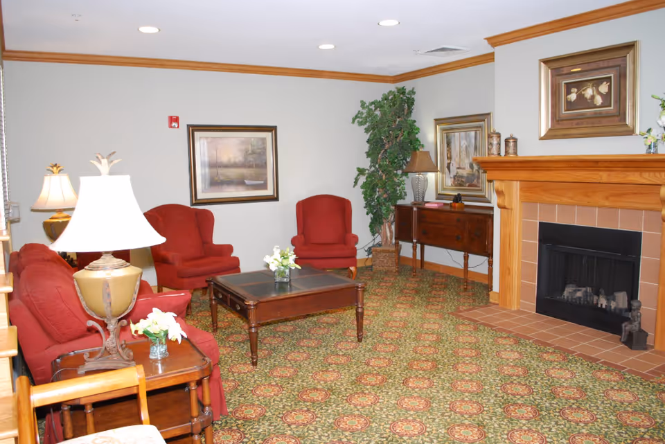 A cozy living room with red upholstered chairs and a sofa arranged around a wooden coffee table. The room features a patterned carpet, a wooden fireplace with a framed painting above it, a side table with a lamp, and framed artwork on the walls. There is also a tall potted plant in the corner.