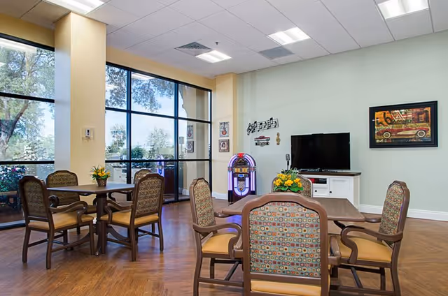 A bright common area with wooden flooring featuring two tables each surrounded by four cushioned chairs with patterned upholstery. Large windows on the left side allow natural light to fill the room, showing trees outside. On the far wall, there is a jukebox, a flat-screen TV on a white cabinet, and framed artwork. The room has a welcoming and comfortable atmosphere.