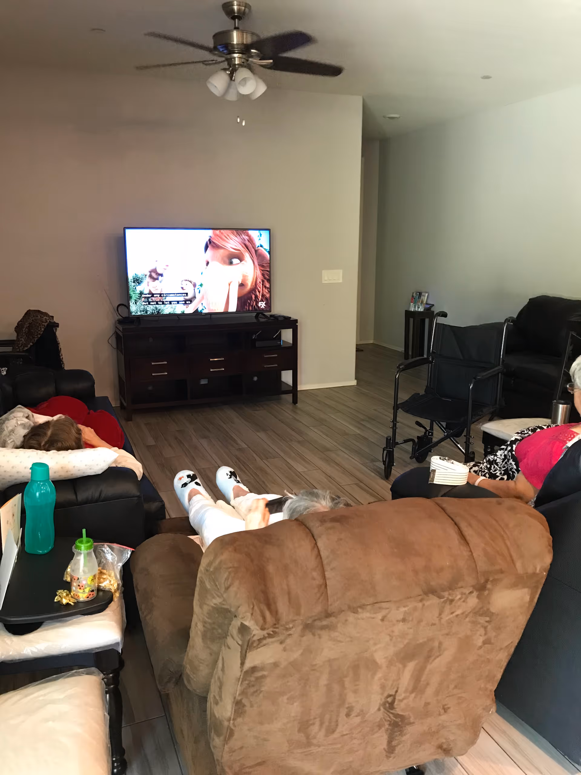 A living room with several people seated on recliners and sofas watching a television mounted on a wooden TV stand. The floor is wooden, and there is a ceiling fan with lights above. A wheelchair is visible near one of the sofas, and a small table with a green water bottle and snacks is in the foreground.