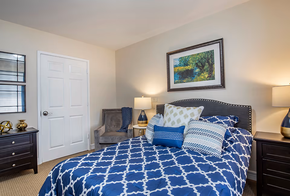 A neatly made bed with a blue and white patterned comforter and multiple decorative pillows in a bedroom. The room features a gray upholstered headboard with nailhead trim, two dark wooden nightstands with lamps, a gray armchair with a blue throw blanket, a white door, and a framed landscape painting on the wall.