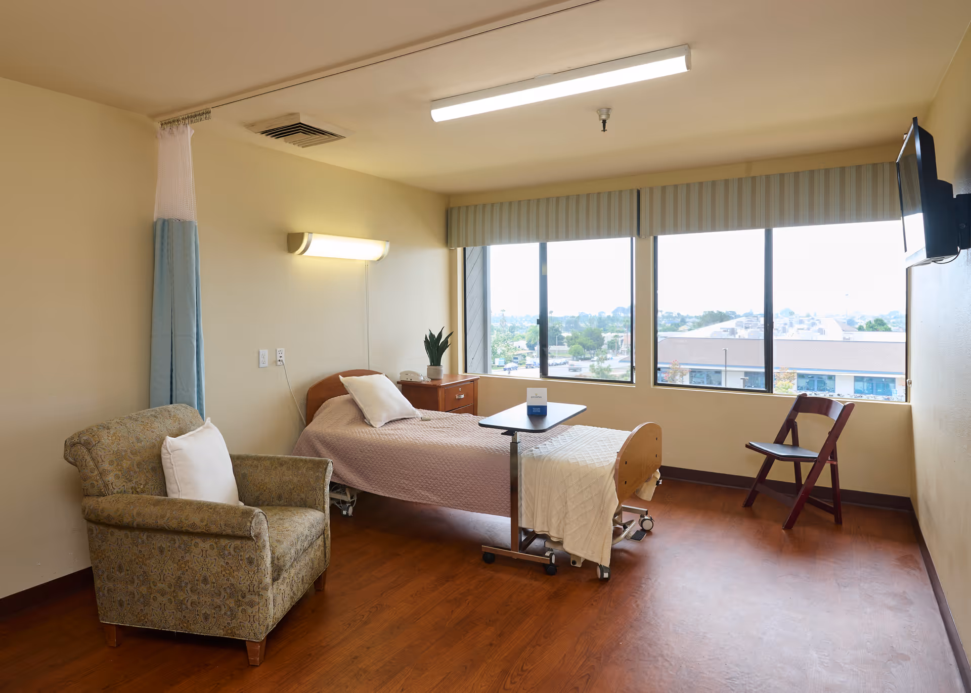 A nursing facility room with a single bed covered in a light brown blanket and a white pillow. Next to the bed is a wooden nightstand with a potted plant and a telephone. A small overbed table is positioned over the bed. There is a patterned armchair with a white pillow on the left side of the room and a folding wooden chair near the large windows that provide a view of buildings and trees outside. The room has wooden flooring, beige walls, a ceiling light, and a wall-mounted TV on the right.