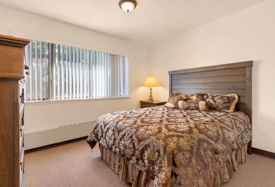 A furnished bedroom with a large bed dressed in a patterned brown comforter, a wooden headboard, nightstand with lamp, dresser, and a wide window with vertical blinds.