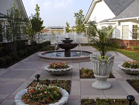 Outdoor courtyard area with a central multi-tiered water fountain surrounded by potted plants and flower beds. The courtyard is paved with large square tiles and bordered by white buildings and a white fence in the background.