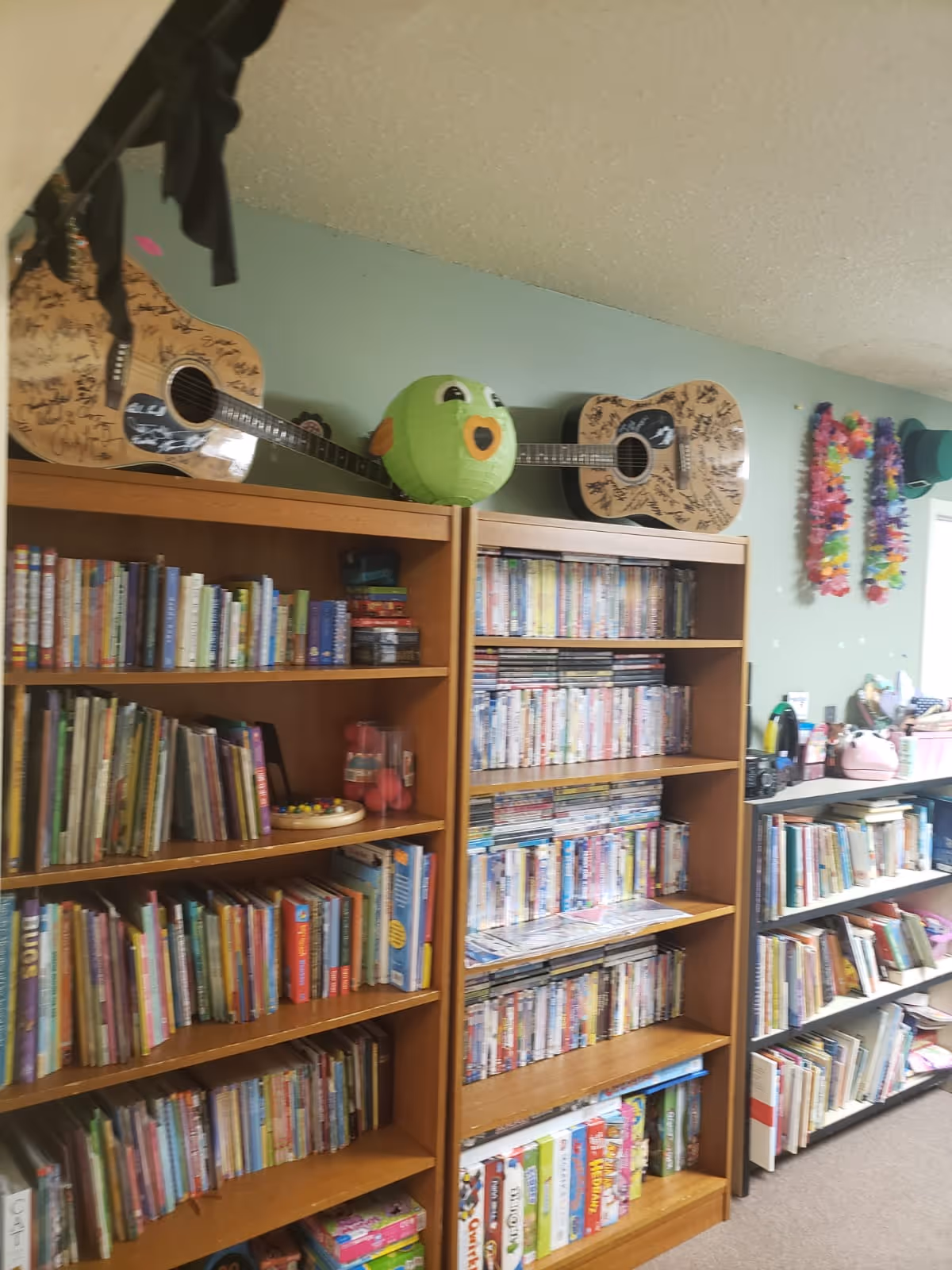 Interior room with wooden bookshelves filled with books, DVDs, and board games. Two guitars with signatures are displayed on top of the shelves along with a green plush toy. Colorful leis and hats are hanging on the wall near a window.