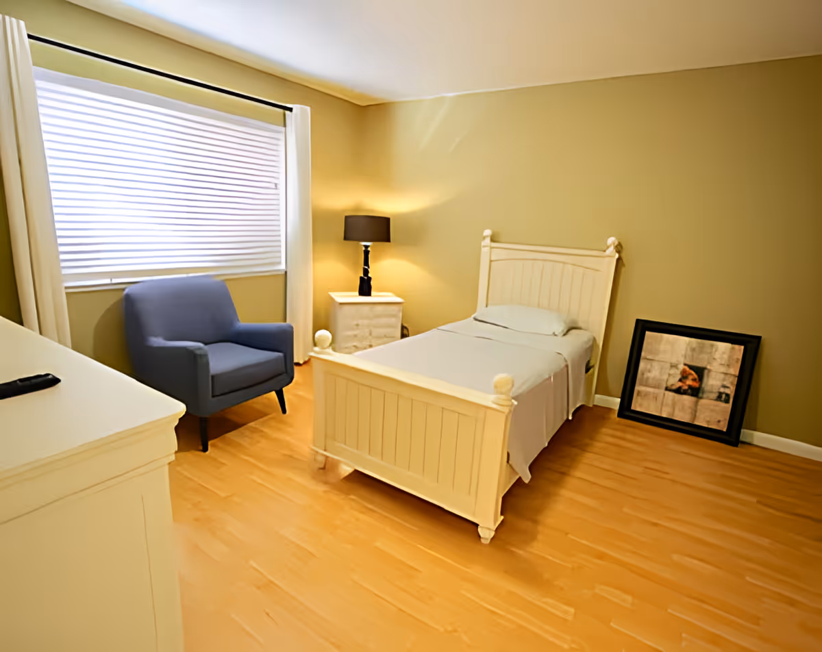 A simple bedroom with a single white wooden bed, a bedside table with a lamp, a blue armchair, a window with white blinds and curtains, and a framed picture resting on the floor against a beige wall.
