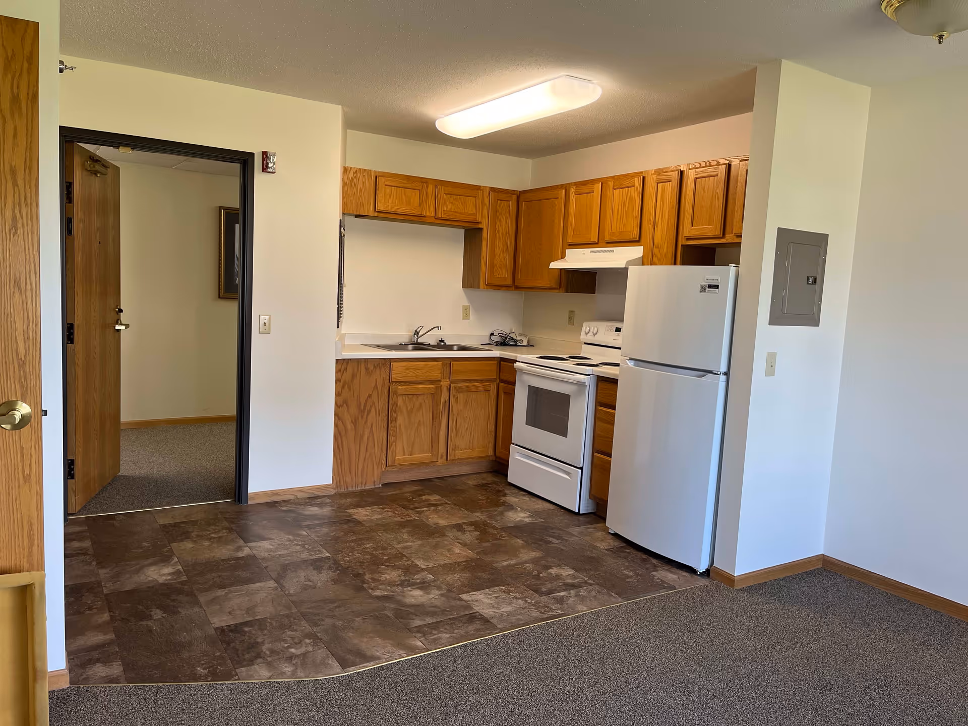 A small kitchen area in a senior living facility with wooden cabinets, a white refrigerator, a white stove with an overhead vent, and a sink. The floor in the kitchen area is covered with brown patterned tiles, while the adjacent area has carpet. There is an open doorway leading to another room with carpet and a wooden door.