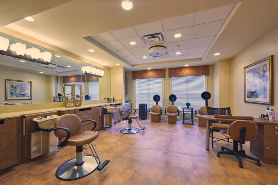 A well-lit salon area in an assisted living facility featuring two brown salon chairs in front of sinks and mirrors on the left side, three brown chairs with hair dryers in front of large windows with blinds, and a manicure table with two chairs on the right side. The room has a warm color scheme with beige walls, a tiled floor, and framed floral artwork on the walls.