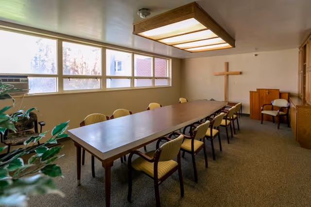 A meeting or conference room with a long rectangular wooden table surrounded by yellow cushioned chairs. The room has a large window letting in natural light, a cross mounted on the wall, a wooden cabinet, and a ceiling light fixture. There is also a plant visible in the foreground.