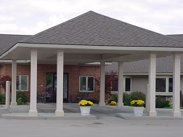 Entrance of a building with a covered driveway supported by white columns, potted yellow flowers on either side, brick walls, and windows in the background.