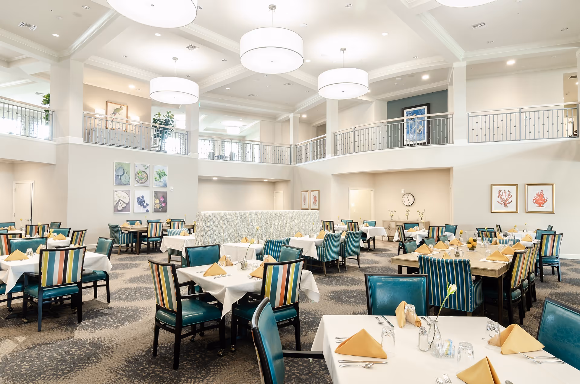A spacious and well-lit dining room with multiple tables covered in white tablecloths, each set with yellow folded napkins, glassware, and silverware. The chairs have teal seats and backs with some featuring colorful striped upholstery. The room has high ceilings with large round pendant lights, a mezzanine level with railings, and walls decorated with framed artwork and plants.