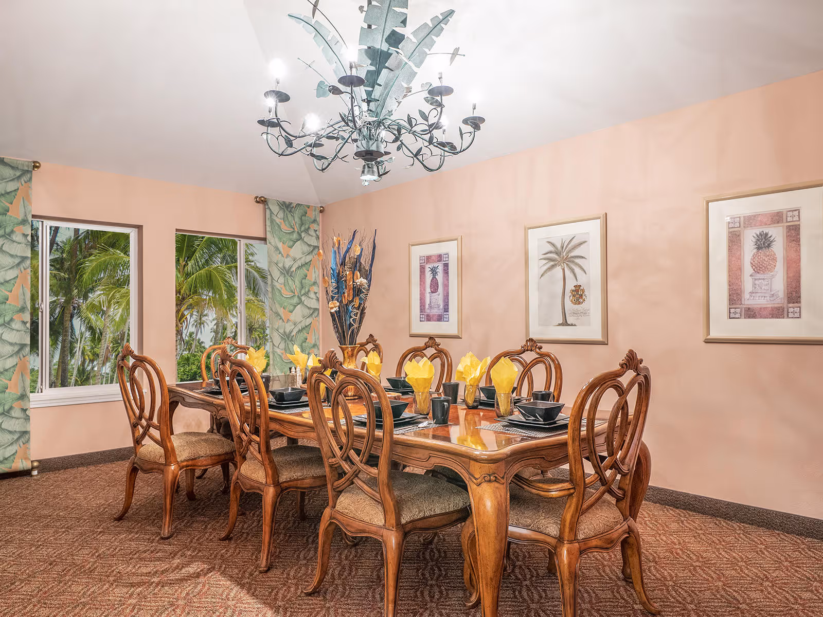 A dining room with a wooden dining table set for eight people. The table has yellow folded napkins in glasses and black dishware. The room features peach-colored walls, three framed botanical prints, patterned curtains with a green leaf design, and a chandelier with leaf-shaped decorations. Two windows show palm trees outside.