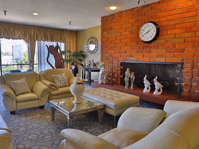 Comfortable seating area with cream leather chairs and an ottoman around a glass coffee table in front of a brick fireplace decorated with figurines and a wall clock.