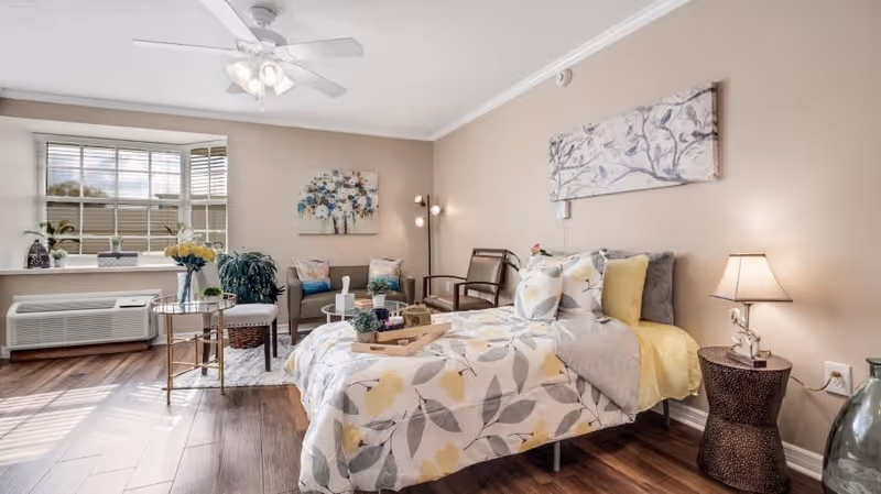 A bright and cozy bedroom with a bed covered in floral patterned bedding in shades of yellow, gray, and white. The room features wooden flooring, a ceiling fan with lights, a window with blinds, and a small seating area with two chairs and a small table. There are decorative plants, a floor lamp, and artwork on the walls, creating a warm and inviting atmosphere.