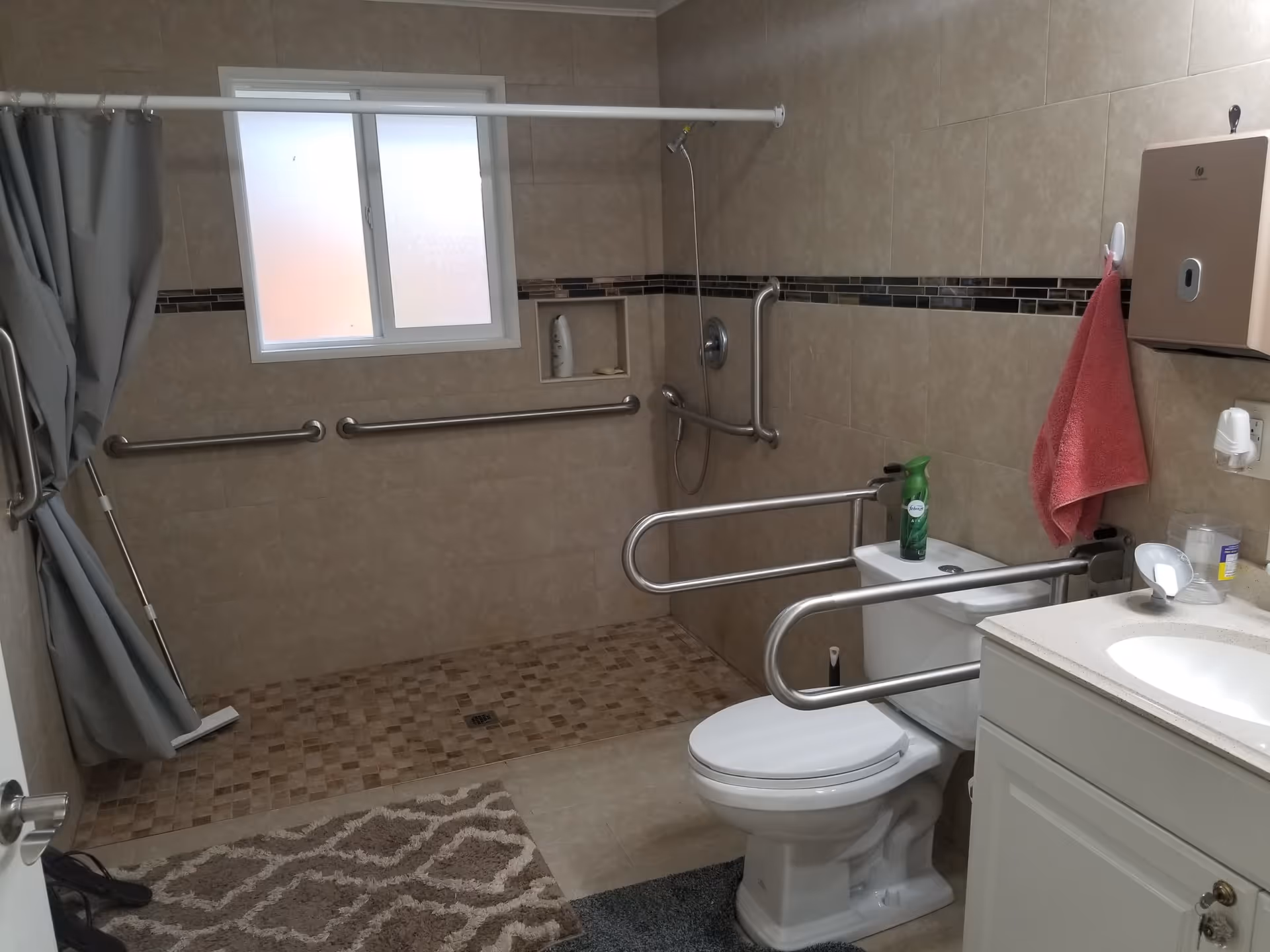 A bathroom with beige tiled walls and floor, featuring a walk-in shower area with grab bars and a shower curtain. There is a white toilet with safety rails on both sides, a white sink with a countertop, a pink towel hanging on the wall, and a window with frosted glass.