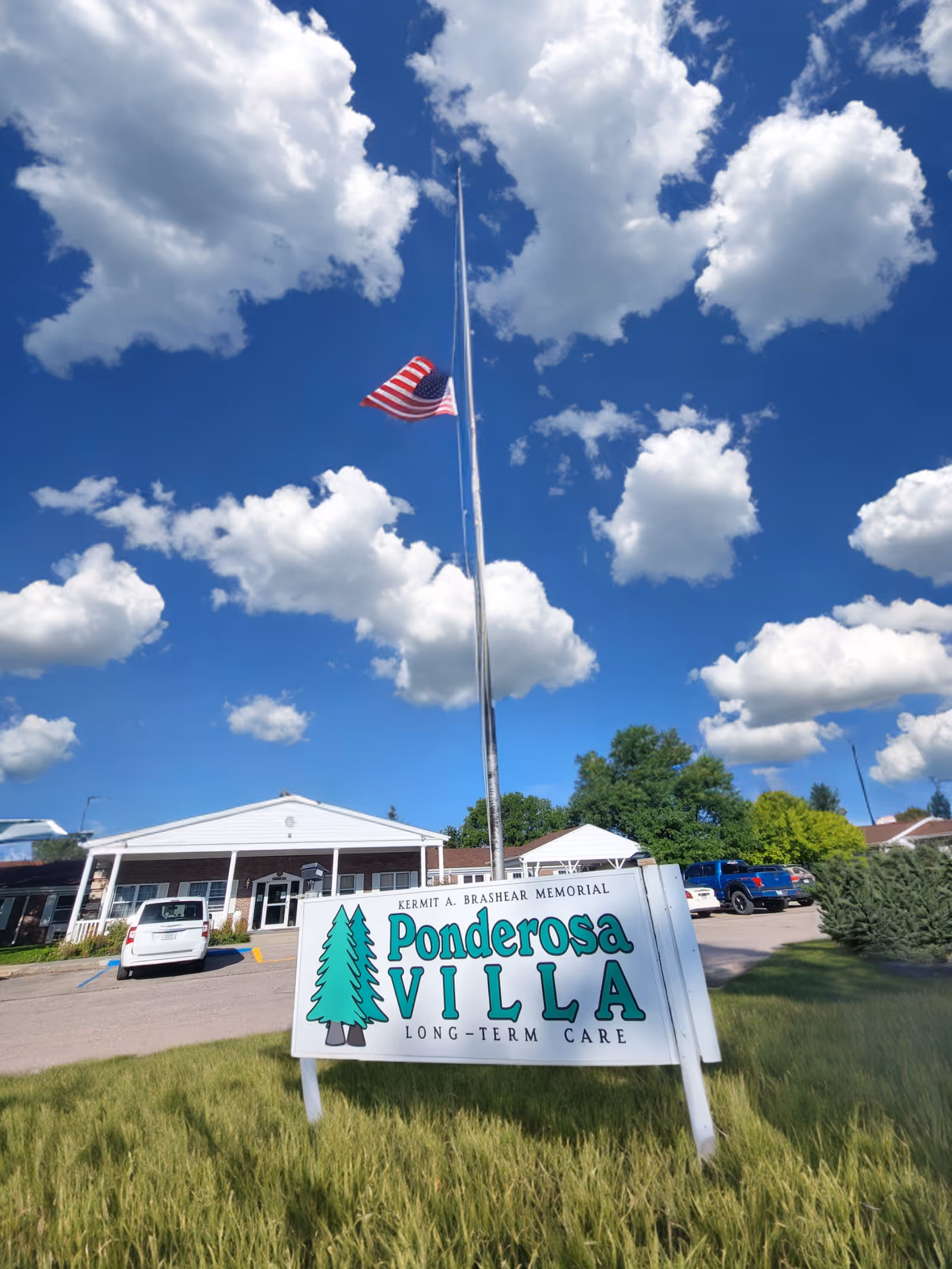 Exterior view of Ponderosa Villa long-term care facility with a white building, parked cars, an American flag at half-mast, and a blue sky with scattered clouds.